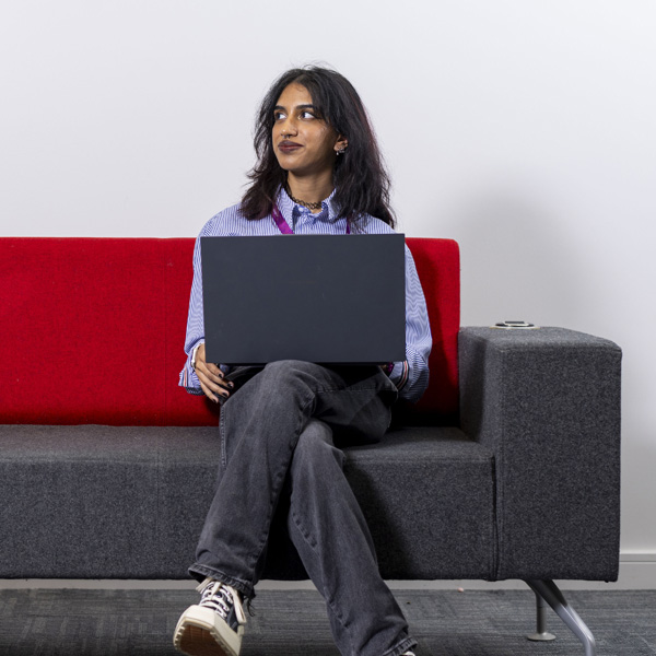 A female student with dark hair wearing a blue shirt is smiling, using her laptop while sitting on an office sofa