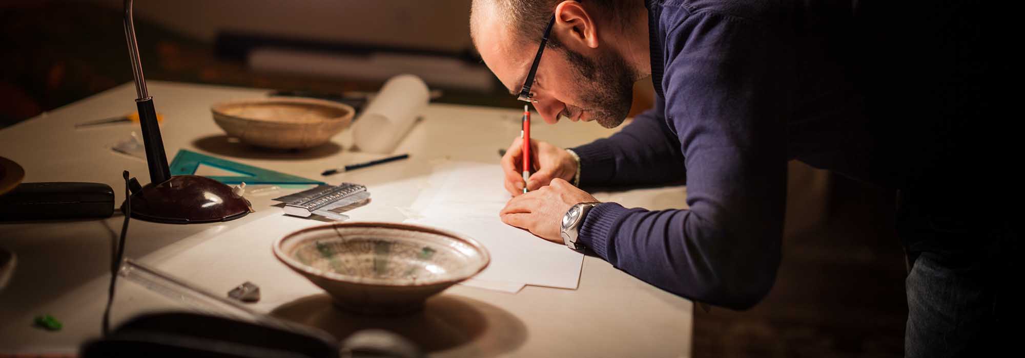 male researcher make notes at a desk, he is surrounded by measuring tools, drafting papers, mechanical pencils and various ceramic bowls which are the object of his research