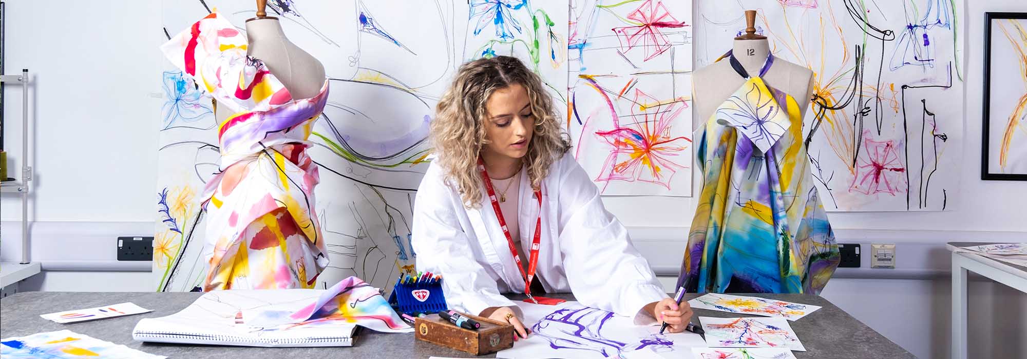 Female student working on a surface pattern design in our art and design studios. Her designs feature bold colours on stark white backdrops. She is in the center of the image with two dressmaker's mannequins either side of her, they are dressed with her u