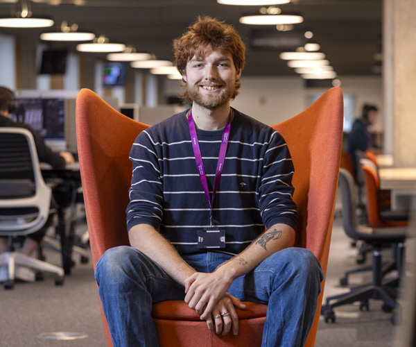 A young male student wearing a striped top and jeans is smiling and sitting in a red chair in an open learning space in the Catalyst building