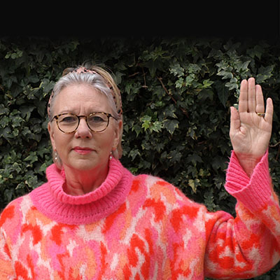 Woman in a bright pink and orange jumper raises one hand against a dark leafy background.