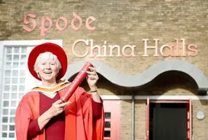 Woman in red academic robes holds a diploma outside the Spode China Halls.
