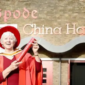 Woman in red academic robes holds a diploma outside the Spode China Halls.