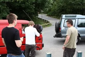 Small film crew records a scene beside red and blue vans on a quiet roadside.