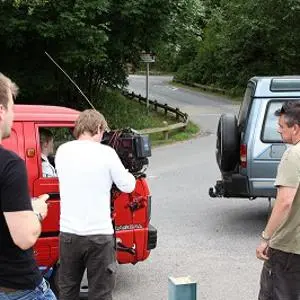 Small film crew records a scene beside red and blue vans on a quiet roadside.