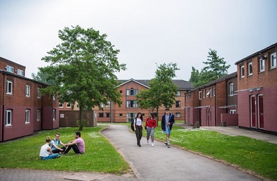 Students walk and sit in a grassy courtyard surrounded by red-brick university residences.