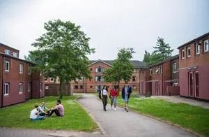 Students walk and sit in a grassy courtyard surrounded by red-brick university residences.