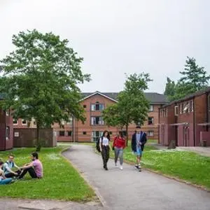 Students walk and sit in a grassy courtyard surrounded by red-brick university residences.