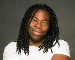 Smiling man with long dreadlocks poses in a white T-shirt against a dark grey background.