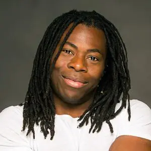 Smiling man with long dreadlocks poses in a white T-shirt against a dark grey background.
