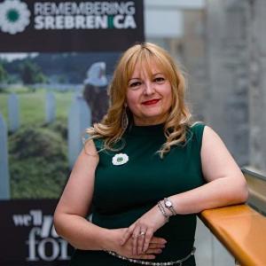 Blonde woman in a green dress stands beside a 'Remembering Srebrenica' display.