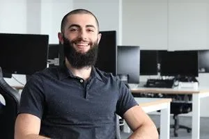 Smiling bearded man sits in a computer lab with rows of monitors behind him.
