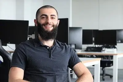 Smiling bearded man sits in a computer lab with rows of monitors behind him.