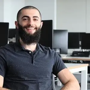 Smiling bearded man sits in a computer lab with rows of monitors behind him.