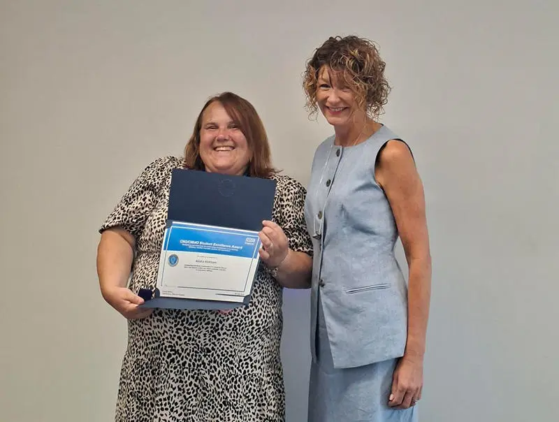 Two smiling women standing together with one holding a framed certificate