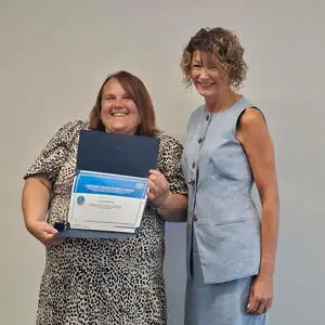 Two smiling women standing together with one holding a framed certificate