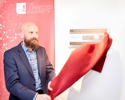 Bald, bearded man unveils a wall display by pulling away a red cloth.