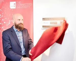Bald, bearded man unveils a wall display by pulling away a red cloth.