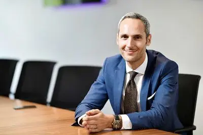 Smiling man in a navy suit sits at a conference table in a modern meeting room.