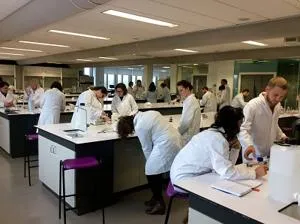 Students in white laboratory coats work at benches in a busy biomedical science lab.