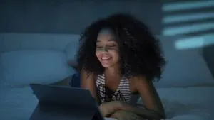Smiling woman uses a tablet while sitting in bed in a dimly lit room.