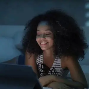 Smiling woman uses a tablet while sitting in bed in a dimly lit room.