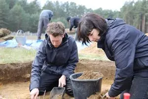 Two archaeologists kneel in an excavation trench and carefully clear soil.