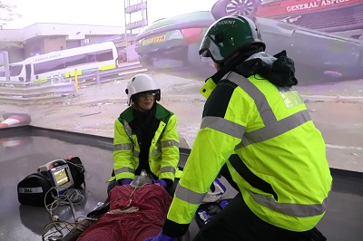 Two people in high vis treat a mock patient in front of a screen depicting a road traffic collision.