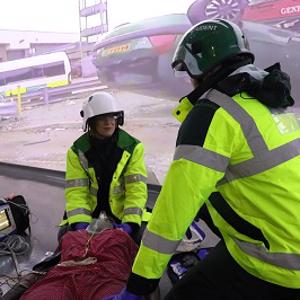 Two people in high vis treat a mock patient in front of a screen depicting a road traffic collision.
