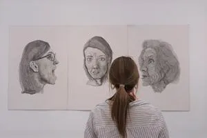 Visitor views three large pencil portraits displayed on a white gallery wall.