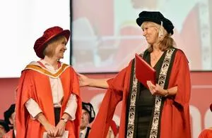 Two women in red academic robes talk to each other during a graduation ceremony.