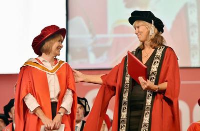 Two women in red academic robes talk to each other during a graduation ceremony.