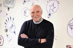 Older man with folded arms smiles in front of a wall covered with colourful idea sketches.