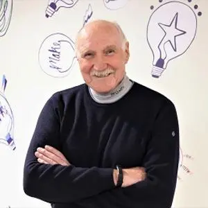 Older man with folded arms smiles in front of a wall covered with colourful idea sketches.