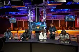 Panel of speakers sits behind a long desk in a blue and purple-lit esports arena.