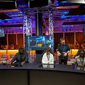 Panel of speakers sits behind a long desk in a blue and purple-lit esports arena.