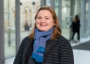 Smiling woman in a blue scarf and striped coat poses in a bright university atrium.