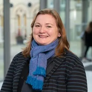 Smiling woman in a blue scarf and striped coat poses in a bright university atrium.