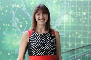 Smiling woman in a black patterned dress stands before a green digital network graphic.