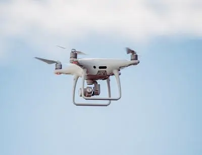 White quadcopter drone hovers against a pale blue, lightly clouded sky.