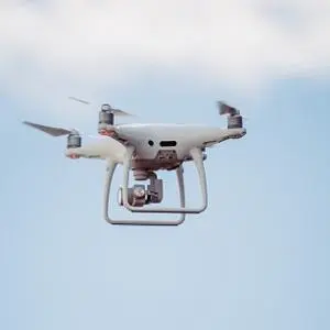 White quadcopter drone hovers against a pale blue, lightly clouded sky.