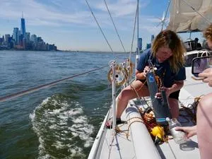 Scientist leans over a sailboat to collect a water sample, with a city skyline behind.