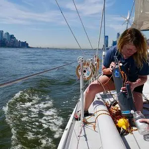 Scientist leans over a sailboat to collect a water sample, with a city skyline behind.