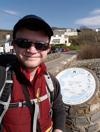 Hiker in sunglasses takes a selfie at a hilltop viewpoint overlooking a coastal village.