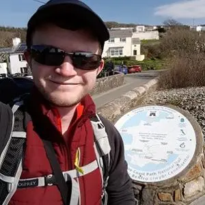 Hiker in sunglasses takes a selfie at a hilltop viewpoint overlooking a coastal village.