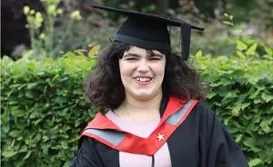 Smiling graduate in cap and black gown with red trim poses outdoors.
