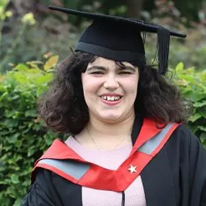 Smiling graduate in cap and black gown with red trim poses outdoors.