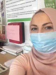 Woman in a pink headscarf and surgical mask takes a selfie in an office.