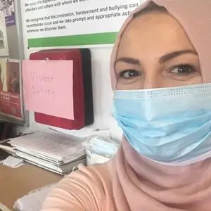 Woman in a pink headscarf and surgical mask takes a selfie in an office.
