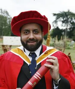 Man in red academic robes holds a diploma outdoors beside trees.